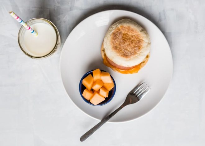 10 Toddler Breakfast Ideas - a photo of a breakfast sandwich next to a blue cup of orange cantelope on a white plate and white background - click photo for full written recipes