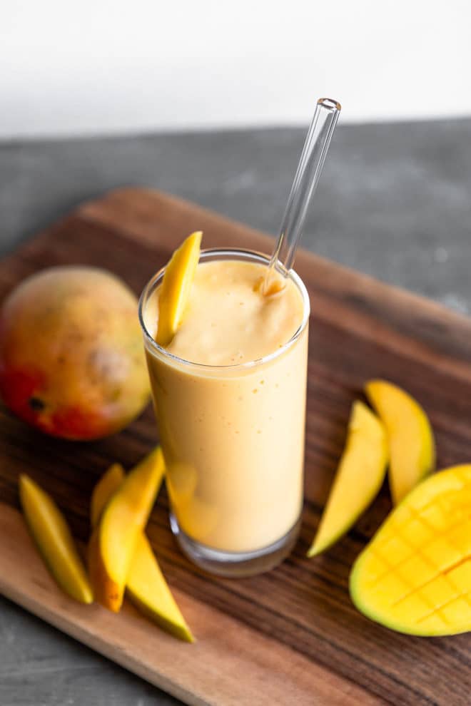 A tropical mango smoothie in a clear glass sitting on a wooden cutting board.