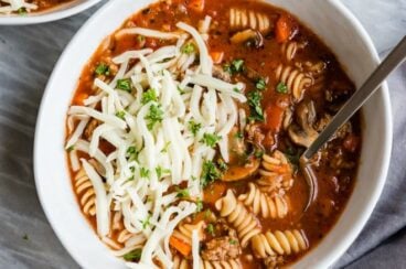 Slow cooker lasagna soup in a white bowl with a spoon and shredded mozzarella cheese.
