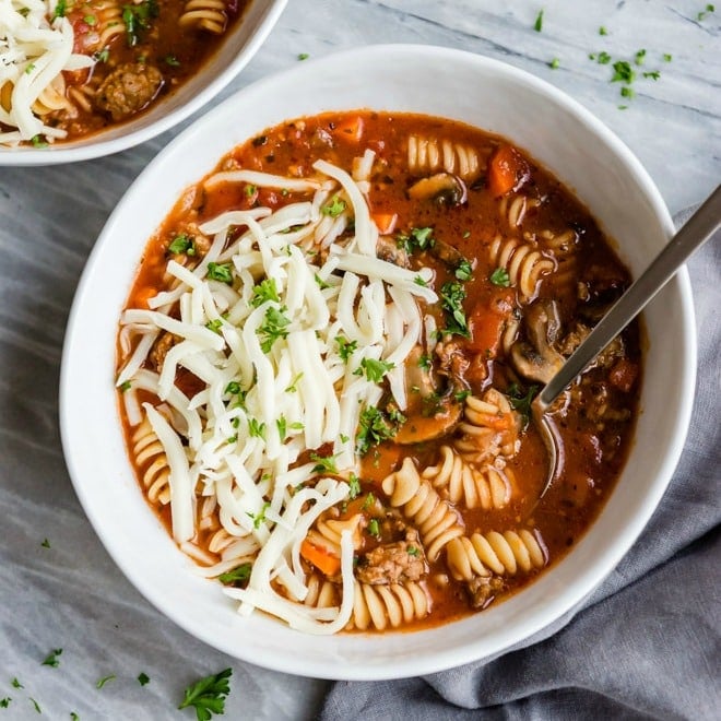 Slow cooker lasagna soup in a white bowl with a spoon and shredded mozzarella cheese.