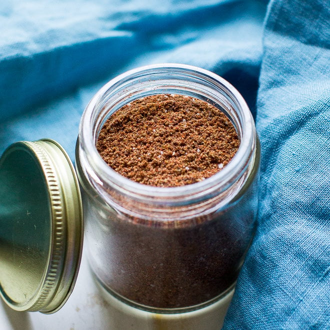 A photo of a clear spice jar with the dark brown taco seasoning. The jar is set upon a blue linen, and the jar's gold lid is next to the jar, leaning against it.
