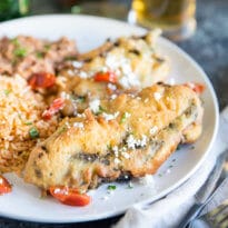A square image of two freshly fried chile rellenos with Mexican Rice and refried beans on a white plate with Dos Equis beer bottle in the background. Half the beer is poured into a glass.