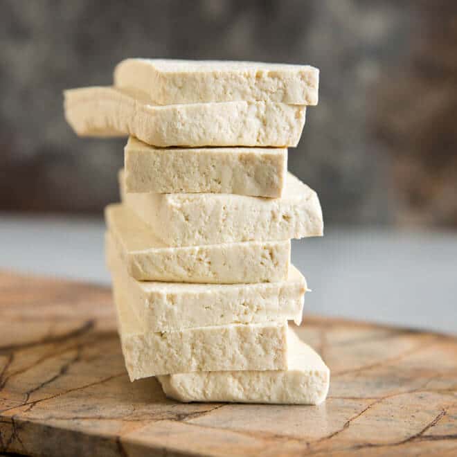 Slices of tofu stacked into a pile on a wood cutting board. 