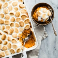 Sweet potato casserole with marshmallows in a white baking dish and a scoop of some in a brown bowl with a fork.