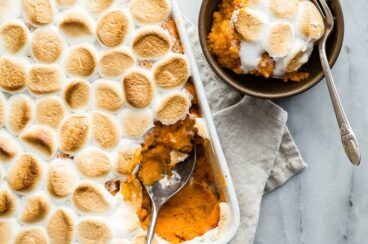 Sweet potato casserole with marshmallows in a white baking dish and a scoop of some in a brown bowl with a fork.