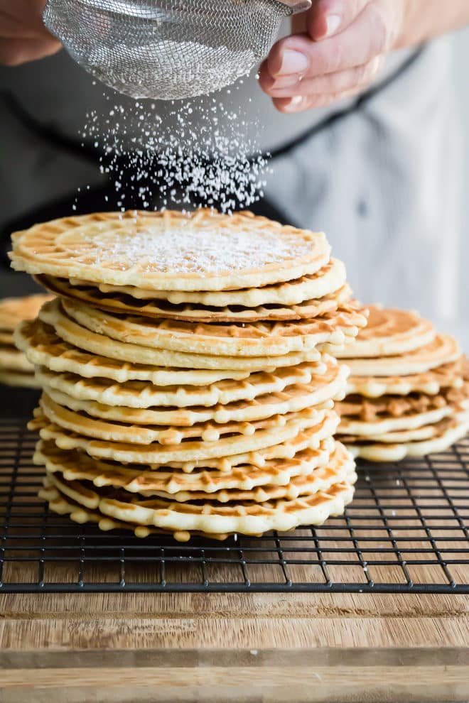 Pizzelle on a cooling rack being dusted with powdered sugar.