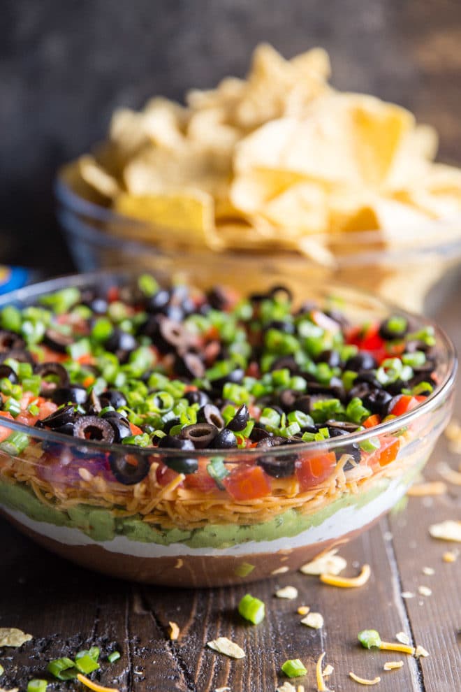 7 Layer Dip in a clear bowl with tortilla chips in the backgound.