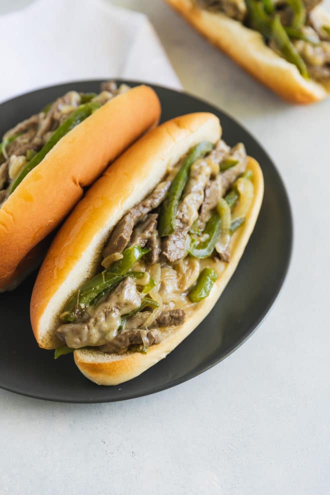 A side shot of two Philly Cheese Steak sandwiches on a round black plate.