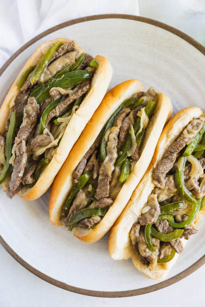 An overhead shot of three Philly Cheese Steak sandwiches on a round white plate.