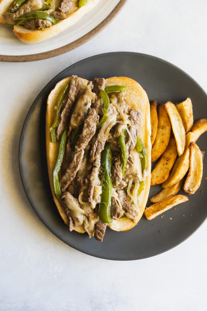 An overhead shot of a Philly Cheese Steak sandwich and steak fries on a round black plate .