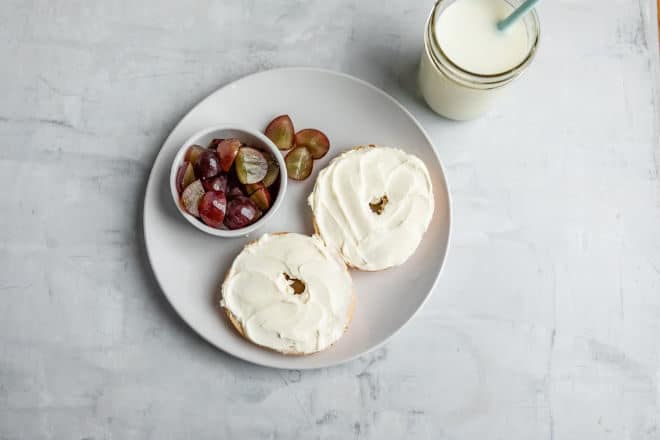 A bagel with cream cheese and a small dish of quartered grapes on a white plate.