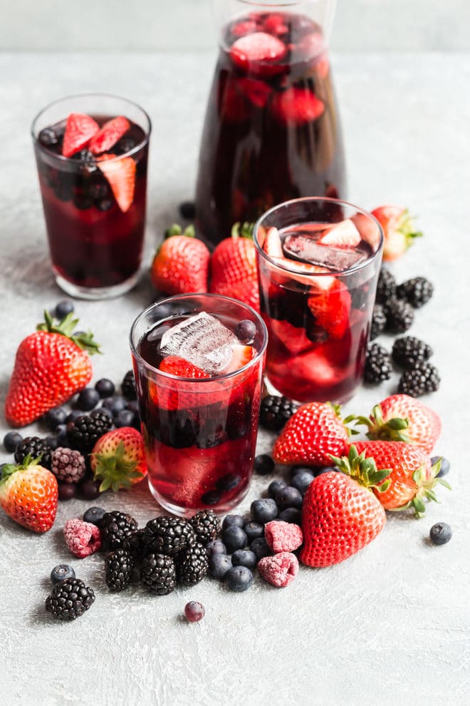 3 glasses and a pitcher of iced tea berry sangria surrounded by piles of fresh berries.