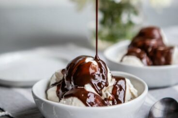 Chocolate sauce being drizzled onto vanilla ice cream in a white bowl.
