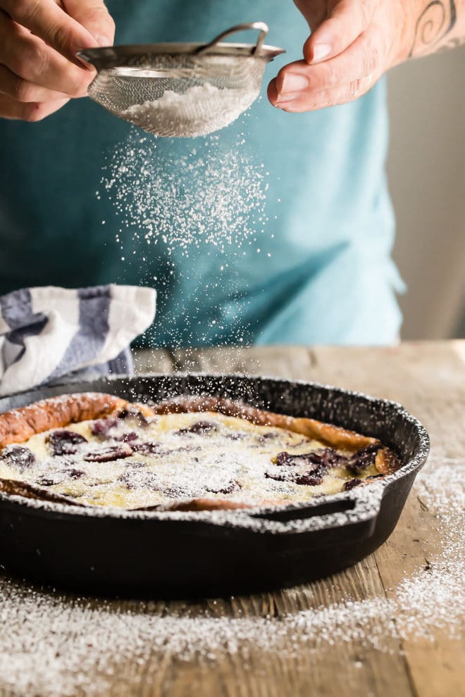 Cherry clafoutis in a black skillet being dusted with powdered sugar.