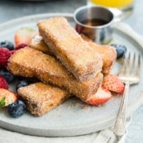 French toast sticks on a white plate with some berries and a side of syrup.