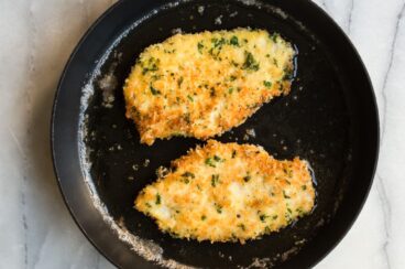 Two pieces of chicken piccata cooking on a black skillet.