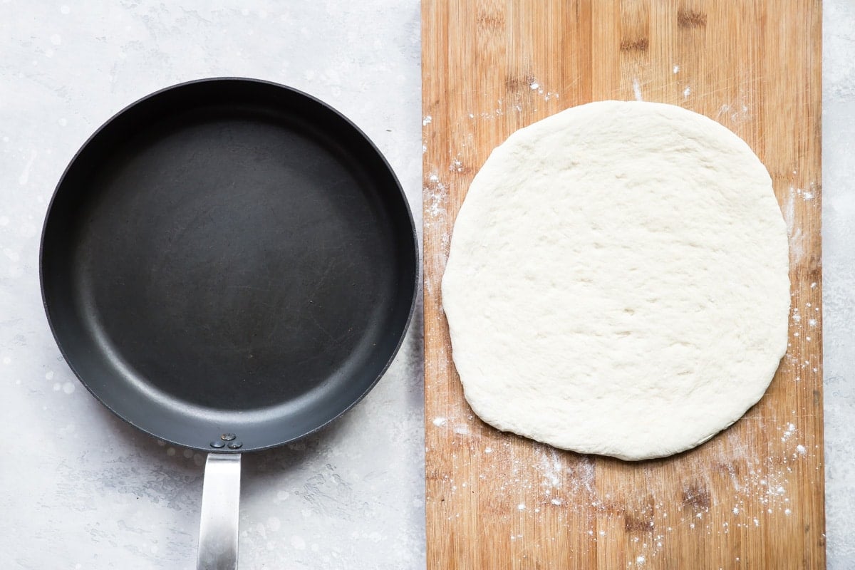 An overhead shot of a cast iron skillet placed next to a circle of dough on a cutting board.