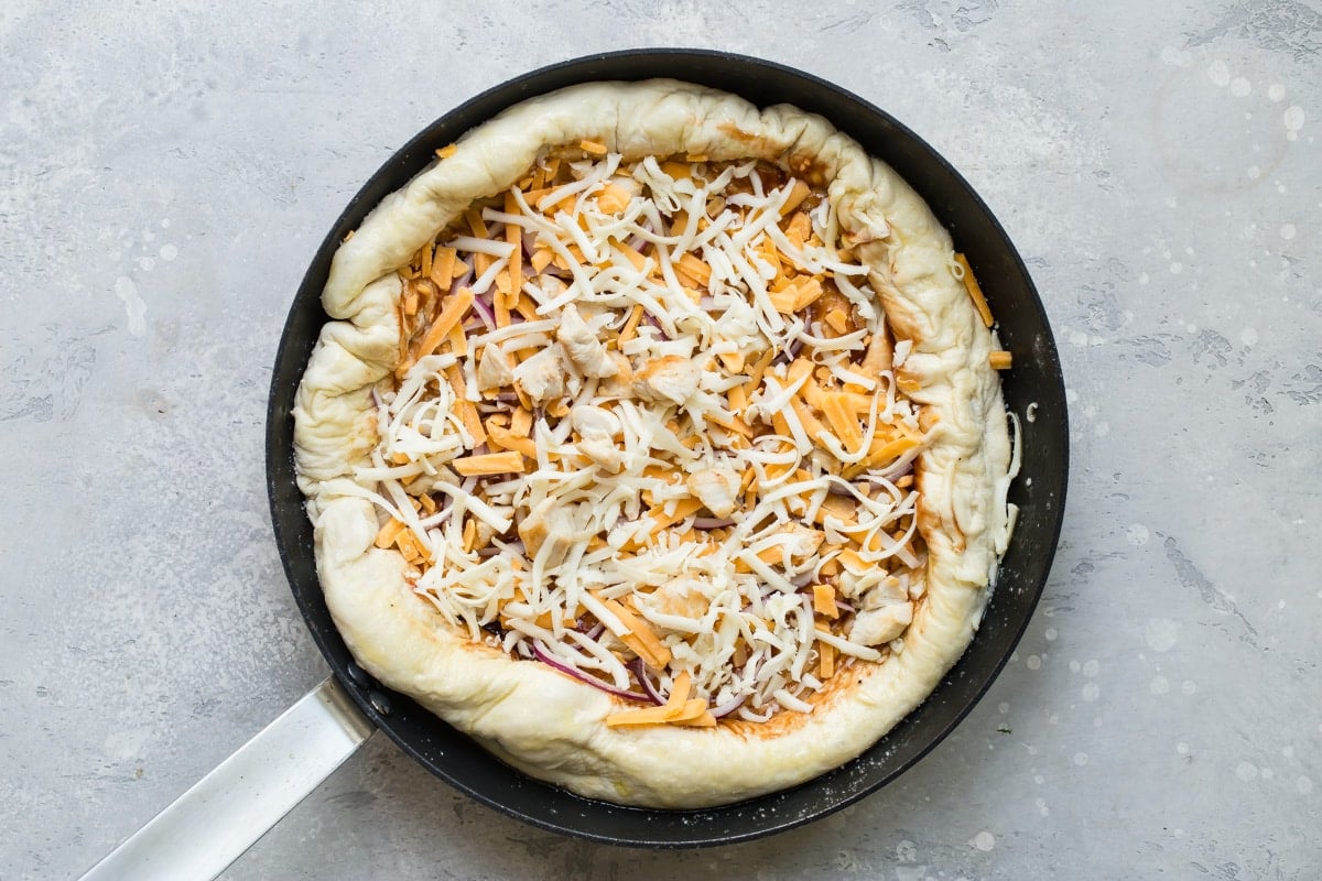 An overhead shot of an uncooked pizza crust with sauce and cheese in a cast iron skillet.