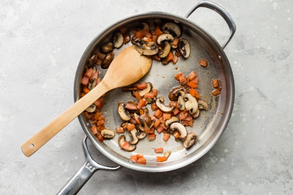 Tomatoes and mushrooms cooking in a silver pan with a wooden spoon.