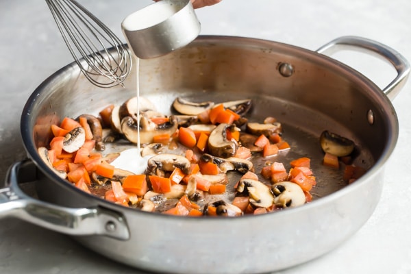 Cream being poured onto cooked mushrooms and tomatoes in a silver pan.