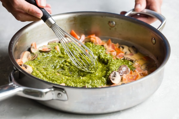 Pesto being stirred into mushrooms and tomato mixture in a silver pan.