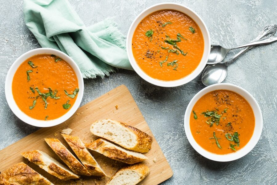 Roasted tomato soup in white bowls with sliced bread on a wood cutting board.
