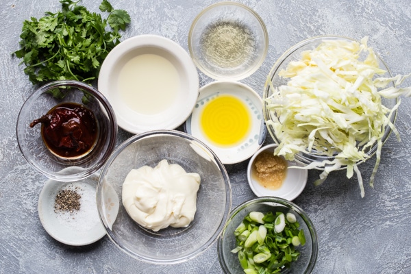 Cabbage slaw ingredients in various bowls on a gray counter top.