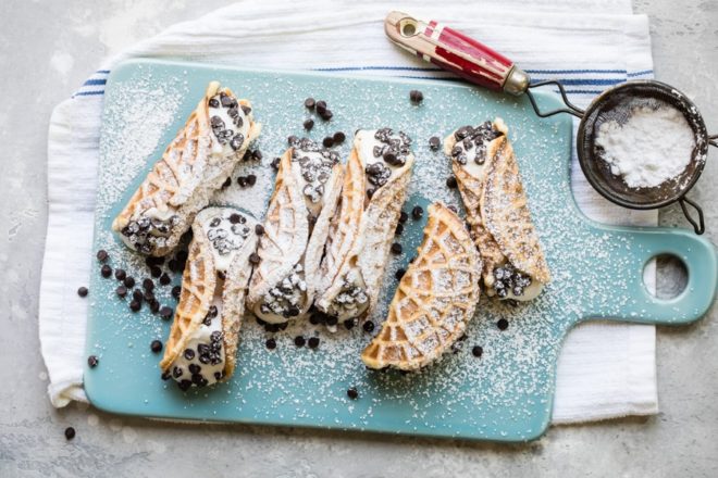 Mock italian cannolis with pizzelle on top of a blue cutting board covered in powdered sugar.