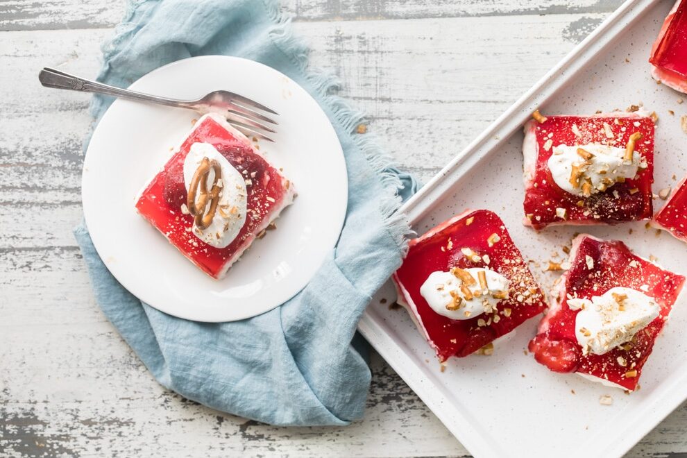 An overhead shot with multiple servings of strawberry pretzel salad on a white serving tray and one serving on a white plate with a silver fork.