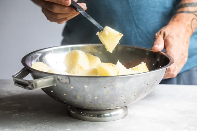 Steamed potatoes in a sliver colander with someone holding a piece of potato on a knife.