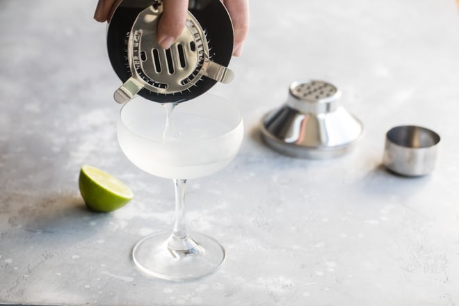 A gimlet being poured from a silver shaker into a clear stemmed glass.