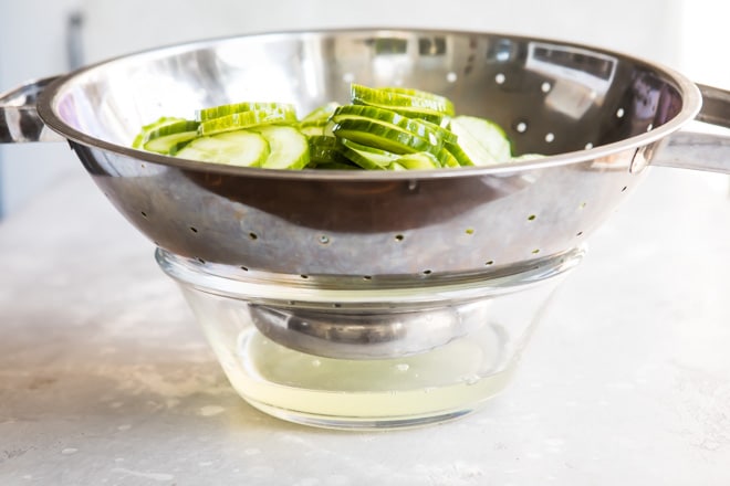 Sliced cucumbers in a silver colander being drained into a clear bowl.