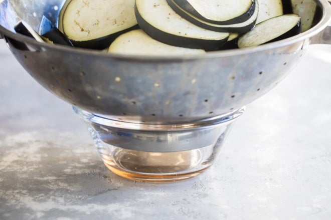 Sliced eggplant in a silver colander being drained into a small clear bowl.