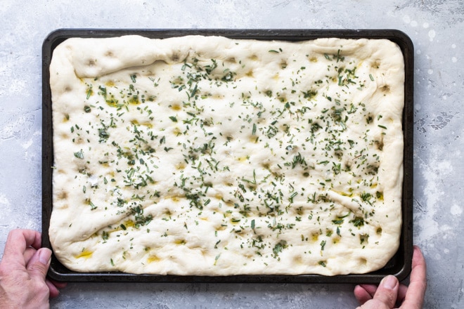 Focaccia dough topped with herbs on a black baking sheet.