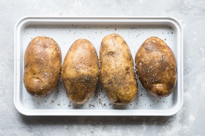 Four whole baked potatoes on a silver baking sheet.