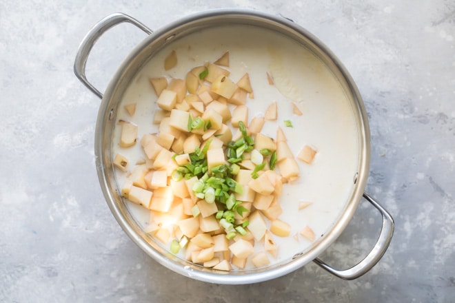 Potatoes and green onion being added to loaded bake potato soup cooking in a silver pot.