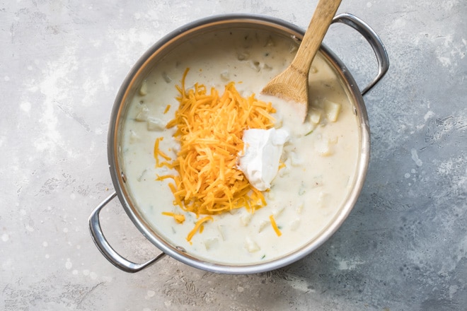 Shredded cheese being added to loaded baked potato soup cooking in a silver pot.