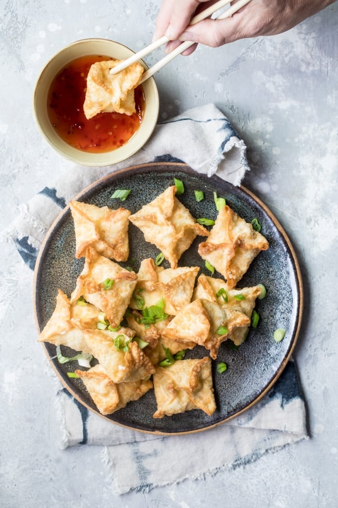 Cream cheese wontons on a plate, garnish with scallions, sweet and sour dipping sauce on the side.