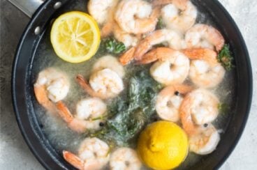 Shrimp being poached in boiling water with lemon halves in a black skillet.
