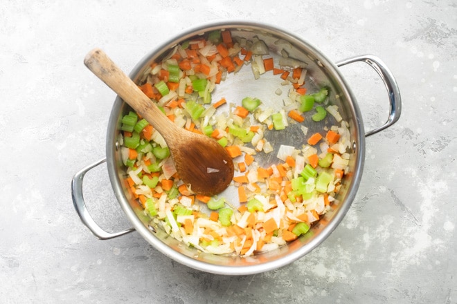Chopped vegetables cooking in a silver pot with a wooden spoon.