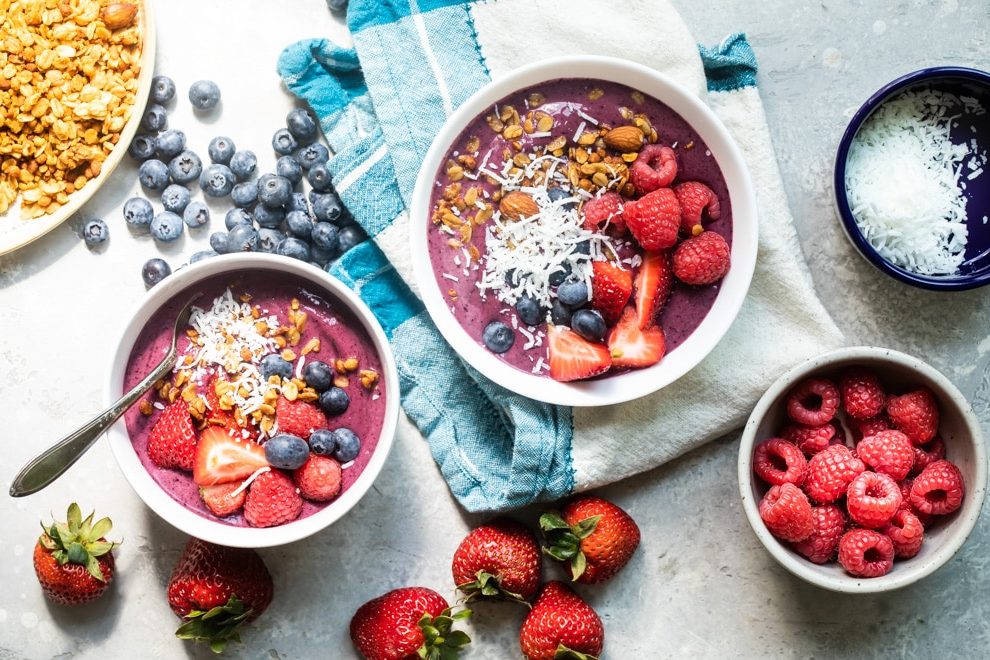 Easy Acai bowl in a white bowl and a larger portion in a white serving bowl surrounded by raspberries, blueberries, and strawberries.