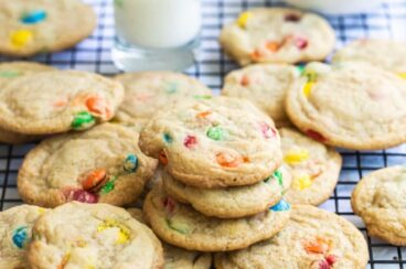 A dozen M&M cookies on a wire rack with a glass of milk and a bowl of M&Ms in the background.