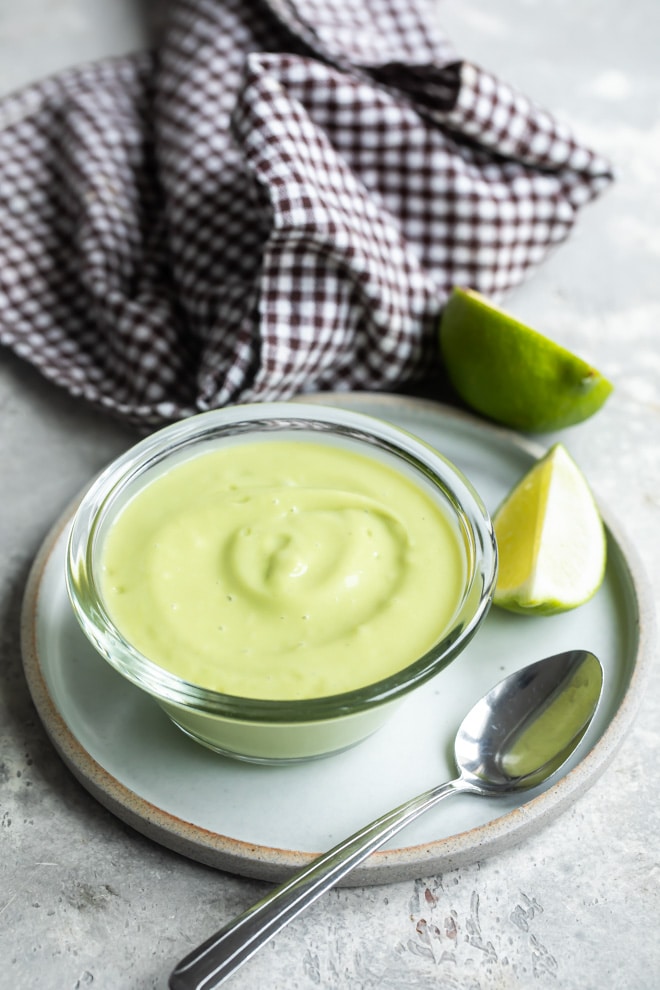 Avocado sauce in a clear bowl on a white plate with a spoon resting on it.
