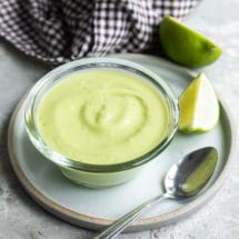 Avocado sauce in a clear bowl on a white plate with a spoon resting on it.