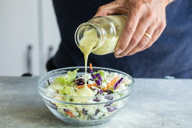 Someone pouring dressing onto coleslaw in a clear bowl.