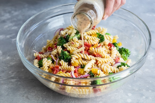 Dressing being poured onto easy cold pasta salad in a clear bowl.