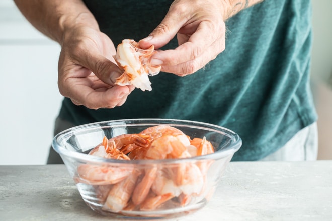 Someone taking the tails off of raw shrimp with some in a clear bowl.