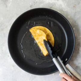 Someone flipping a corn tortilla in a black skillet with tongs.