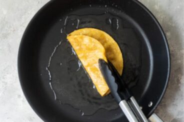 Someone flipping a corn tortilla in a black skillet with tongs.
