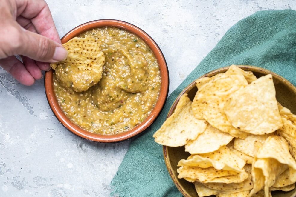 Someone scooping chipotle tomatillo green chili salsa onto a chip and a bowl of chips.